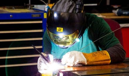 Welder wearing a protective helmet and gloves, focused on welding a metal piece with bright sparks visible.