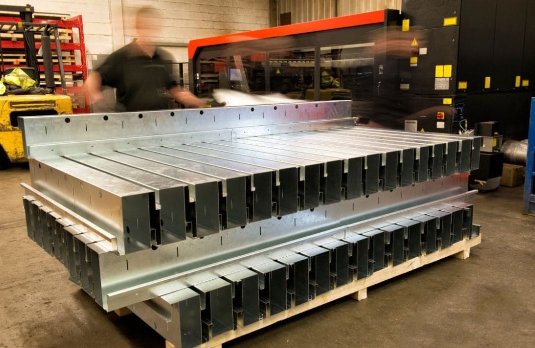 Stacked metal components on a pallet in a workshop, ready for assembly or further processing. Workers move in the background, creating a sense of busy industrial activity.