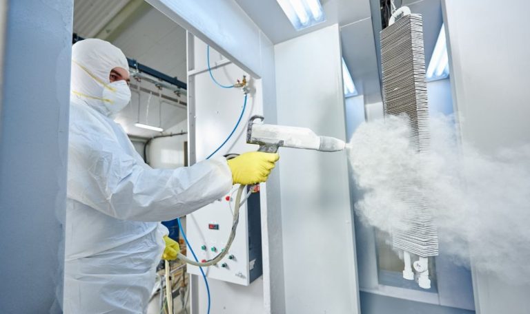 A person in protective gear, including a white suit, face mask, and yellow gloves, operates a powder coating spray gun. The gun releases a thick cloud of powder onto a metal object hanging vertically in a controlled environment with bright overhead lights. The room appears to be a powder coating booth with panels and equipment in the background, indicating an industrial setting.