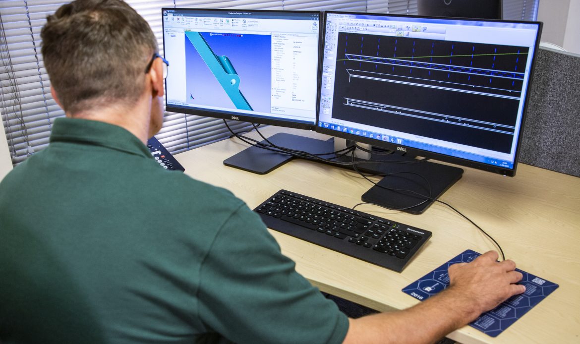 Man using a dual-monitor workstation with CAD software; left screen shows a 3D part model, right screen displays a technical drawing or blueprint in a black-background CAD environment.