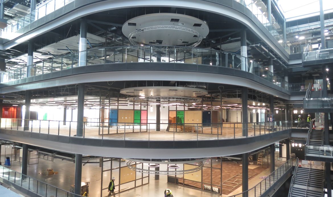 Interior view of a modern multi-story commercial building under construction, with curved balconies, glass railings, open floors, and workers wearing safety gear.