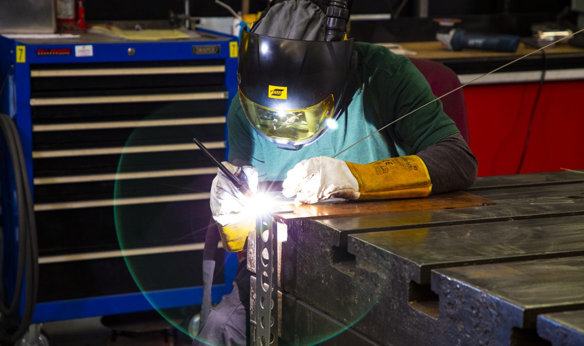 A welder wearing protective gear, including a helmet and gloves, performs TIG welding on a metal frame, creating a bright arc of light.