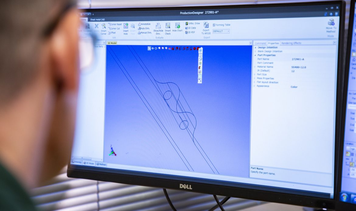 Person using CAD software on a Dell monitor to design a 3D model of a mechanical part, with a detailed wireframe drawing displayed on a blue background.