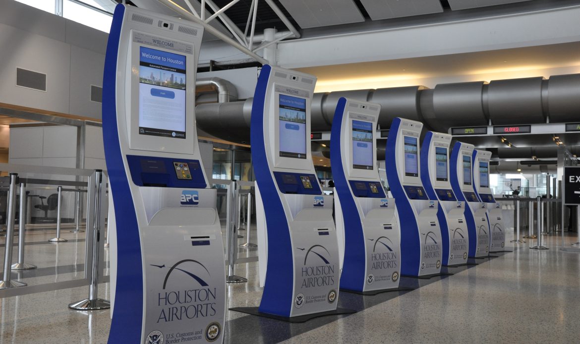 Row of self-service immigration kiosks at Houston Airport, labeled “APC” and “Houston Airports,” with touchscreens displaying a “Welcome to Houston” message.