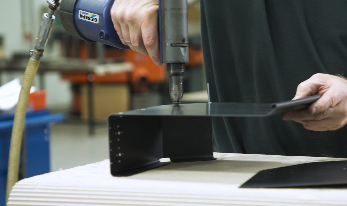 Close-up of a person using a pneumatic rivet gun to fasten a black metal bracket in a workshop setting.