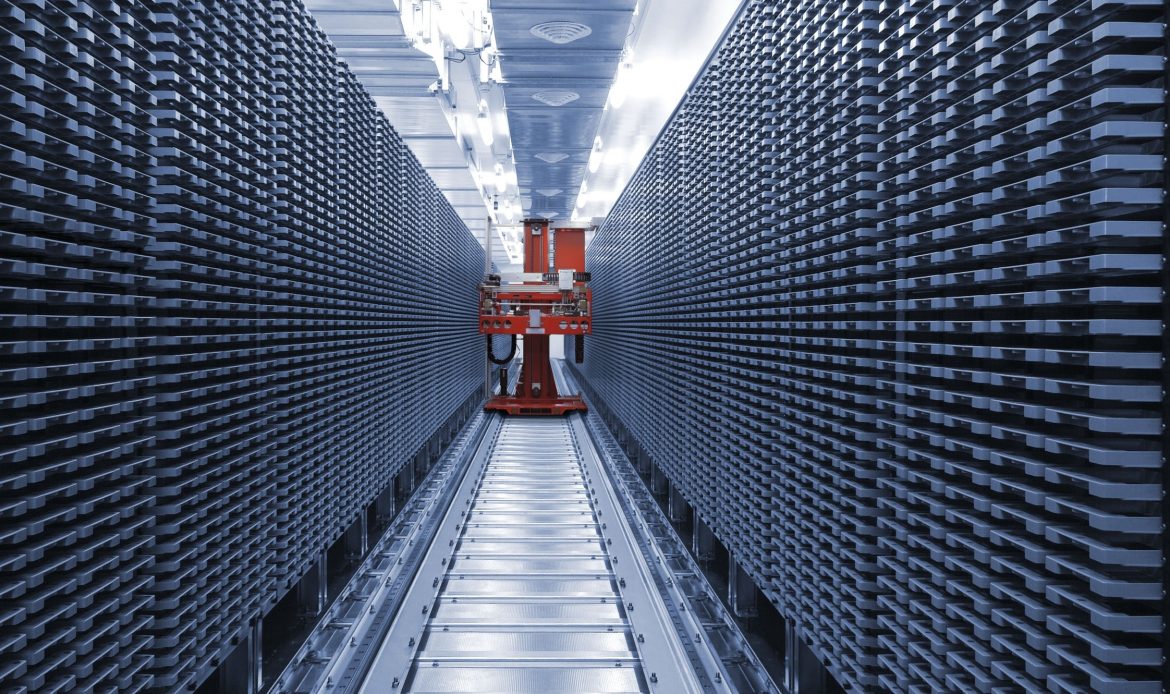 Automated storage and retrieval system with a red robotic arm moving along a track between tall, dense racks of metal trays.