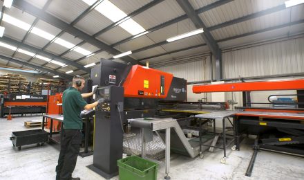 Factory worker operating a large red and black AMADA punching machine in a well-lit industrial workshop with metal sheets and stacked materials in the background.