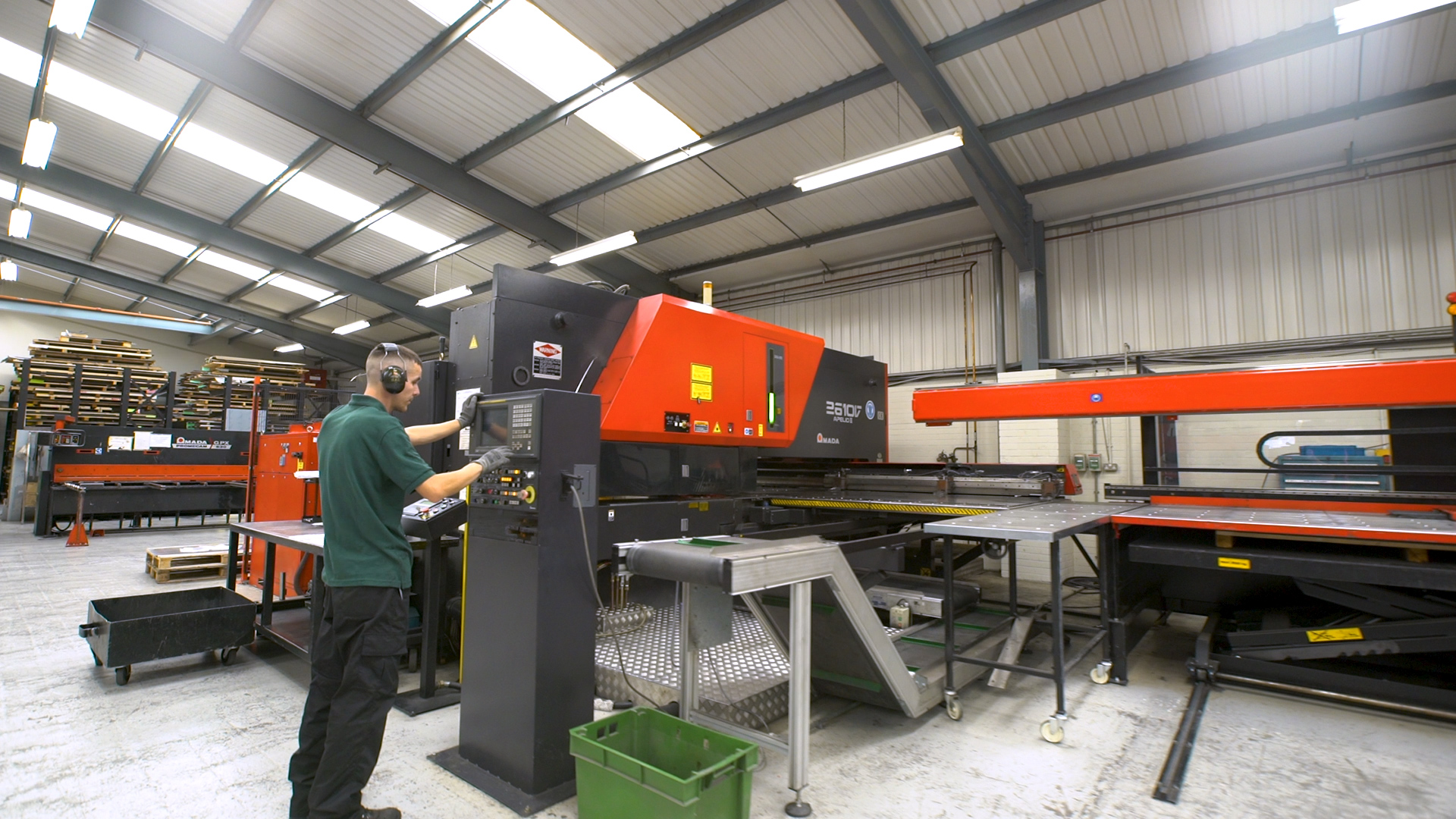 Factory worker operating a large red and black AMADA punching machine in a well-lit industrial workshop with metal sheets and stacked materials in the background.