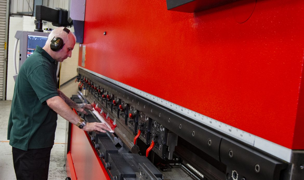 A worker in ear protection operates a large red CNC press brake machine, positioning a metal sheet for bending.