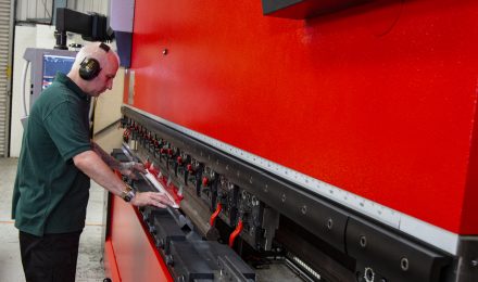 A worker in ear protection operates a large red CNC press brake machine, positioning a metal sheet for bending.