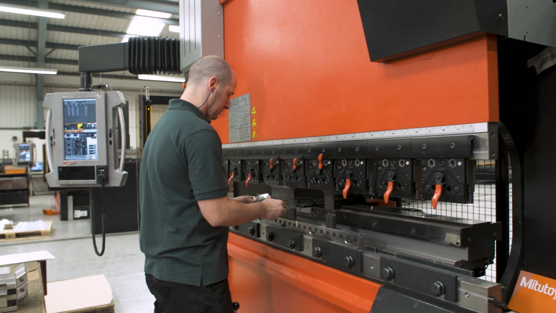 A technician operates a CNC press brake machine, wearing ear protection and inspecting a metal component, with the machine’s control panel and tooling system clearly visible.